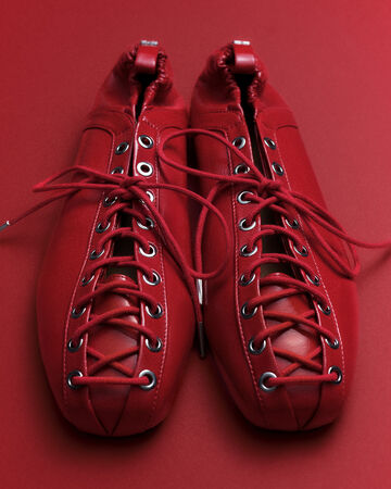 Top-down view of a pair of red GANNI ballerina flats with red lace-up detailing and metal eyelets and elasticated heels on a red background.