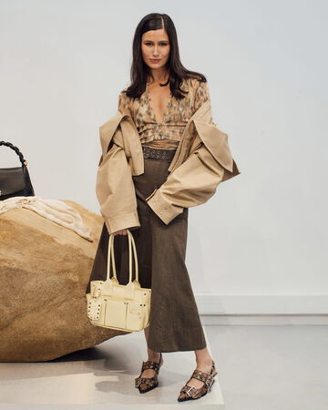Model wearing beige cropped jacket and brown skirt holding a yellow handbag