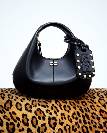 Close-up of the GANNI Small Bou Bag in black leather featuring gold-tone hardware, studded butterfly logo tag, and a top handle, resting on a leopard print background