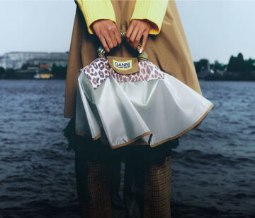 Model standing by water holding a bag with a GANNI x Hunter bag poncho over